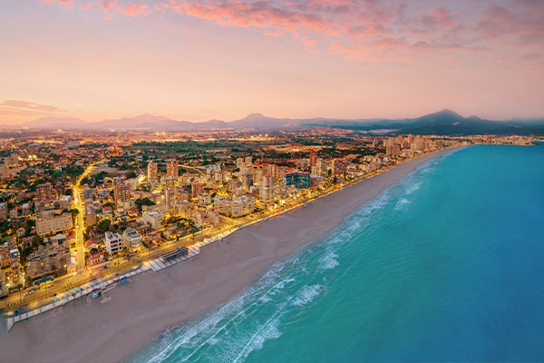 Long stretch of white-sand beach at dusk with illuminated Alicante cityscape to the left and blue Mediterranean waters to the right