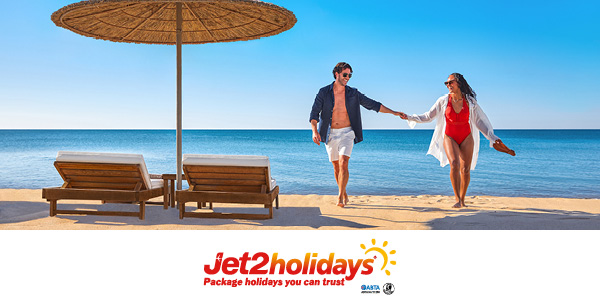 A man and woman holding hand on a beach next to two deckchairs and a parasol