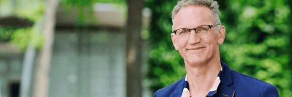 Portret van Michel van Smoorenburg. Michel poseert buiten in een blauw colbertje en een roze blauw gestreept overhemd.