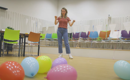 Een vrouw staat in een lege zaal met allemaal gekleurde ballonnen op de grond. Op de achtergrond staan allemaal gekleurde stoelen.