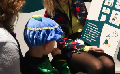 Een kind met een zwarte pietenmuts kijkt naar een plaat met tekst erop die omhoog wordt gehouden door een vrouw. Achter hem staat een andere vrouw.
