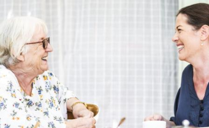Een oudere en jongere vrouw zitten tegenover elkaar en lachen elkaar toe.