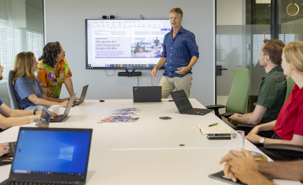 Een aantal Movisiemedewerkers zitten om een tafel en kijken hoe iemand een presentatie geeft.