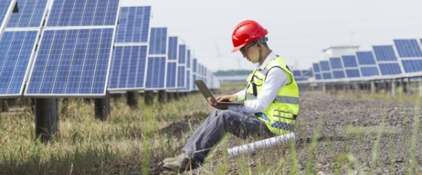 Man zit met laptop op schoot in een veld met zonnepanelen.