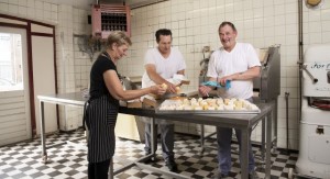 Twee mannen en een vrouw staan in de keuken van een bakkerij en pakken gebakjes in. 