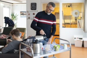 Een persoonlijk begeleider staat bij een serveerwagen in een verzorgingshuis en schenkt iets in een glas uit een van de thermoskannen. 