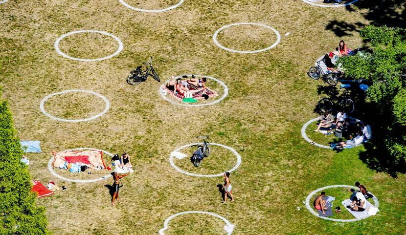 Een bovenaanzicht van een park met cirkels waar groepen mensen op afstand van elkaar zitten en liggen