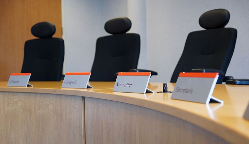 Drie stoelen achter een bureau in de zittingszaal van het College voor de Rechten van de Mens