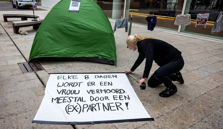 Dolle Mina’s voeren actie tegen het tekort aan opvangplekken voor vrouwen die vluchten voor huiselijk geweld. Een vrouw knielt op de stoep en plakt een groot spandoek vast waarop staat: “Elke 8 dagen wordt er een vrouw vermoord. Meestal door een (ex-)partner!”. Achter haar staat een groen tentje met een papier erop, en aan een waslijn hangen enkele kledingstukken.