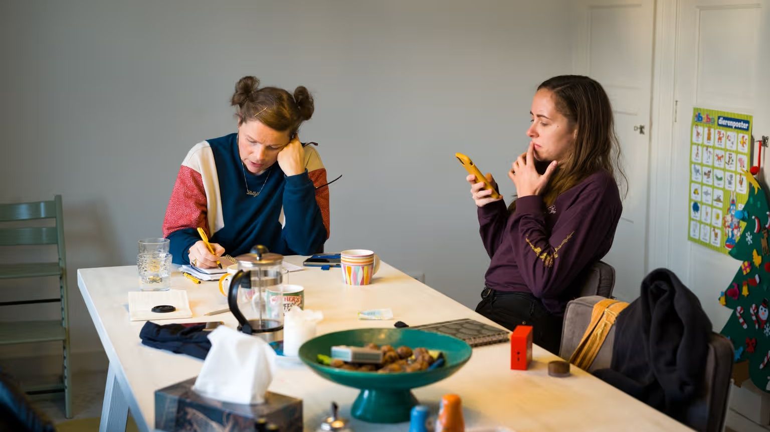 Twee personen aan tafel. Een persoon schrijft, een ander persoon is bezig op haar telefoon