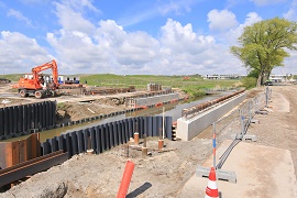 Afbeelding van werkzaamheden om een brug te bouwen. U ziet stalen damwanden en een aantal betonnen wanden, deze liggen aan twee kanten van een tocht.
