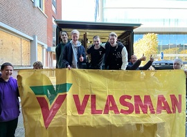 Groep mensen voor een open vrachtwagen en een oud gebouw. Ze houden een gele banner vast met daarop de tekst: Vlasman.
