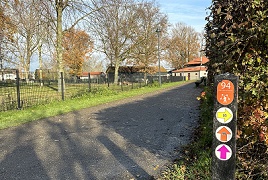 wandelpad met een paaltje met gekleurde pijlen. Op de achtergrond groen en een kinderboerderij.