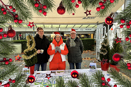 3 mensen achter een marktkraam. Met kerstmutsen op en een warme drank in hun handen.