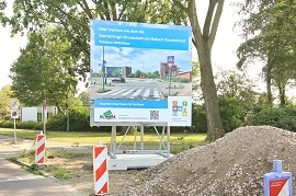 Bouwbord op een straat. Voor het bord een hoop grond en rood/wit gestreepte paaltjes,