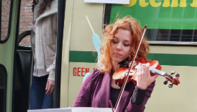 Een vrouw met rood haar en een paars jasje speelt viool van een lessenaar met bladmuziek. Ze zit voor een stemlokaal in een oude bus.