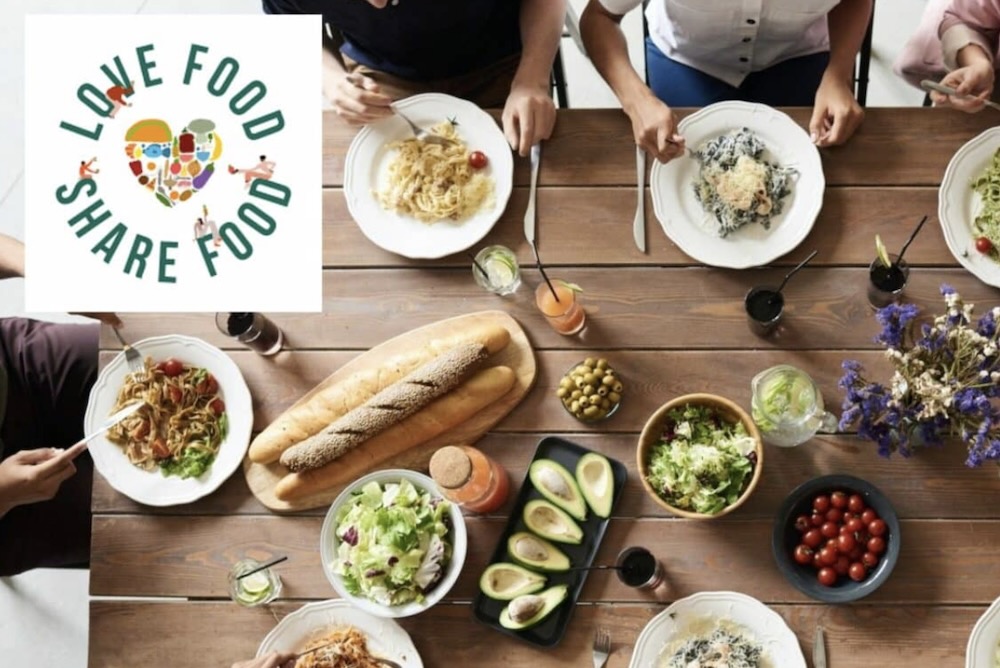 LOVE FOOD SHARE FOOD table photographed from above.  We see bread and vegetables and salad and the hands of people holding cutlery