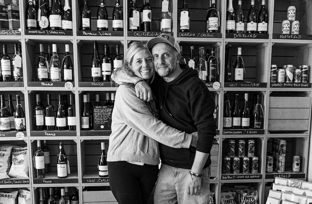 A man and a woman hugging, facing the camera in front of shelves filled with bottles of wine