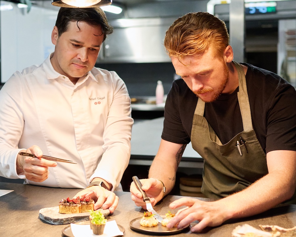 Two men standing in a kitchen with 3 plates of ornate food in front of them. One is holding tweezers