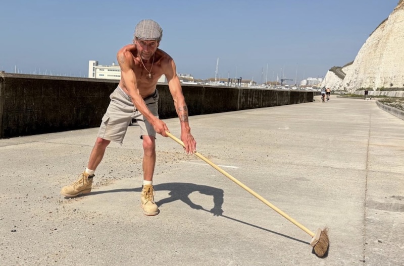 Shirtless man in work boots, shorts and flat cap bending over broom on concrete path under white cliffs