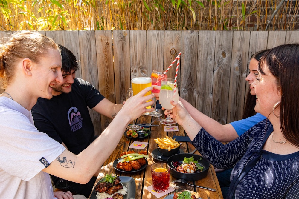 4 people round a table next to a wooden fence holding up glasses as if they are saying cheers. everyone looks happy. The table has lots of food on it