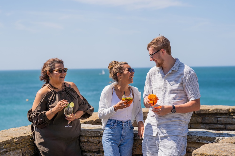 Two women and a man leaning against a wall with sea in the background. It's sunny, they are holding drinks and everyone is smiling