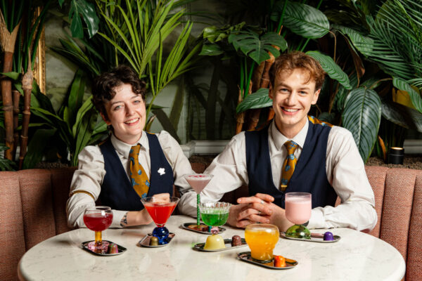 Two smiling people sitting at a table with lots of cocktails in front of them