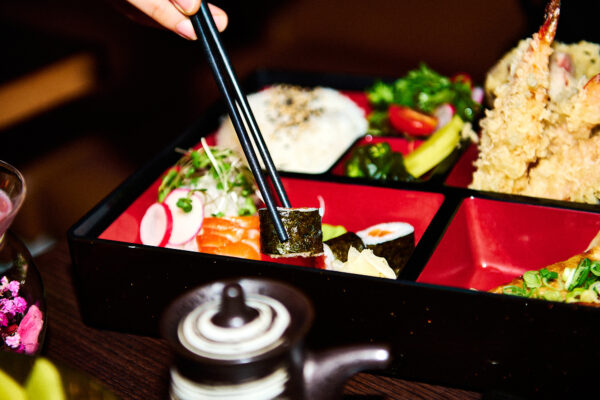 black chop sticks picking up seaweed wrapped sushi roll from a black bento box with red interior sections
