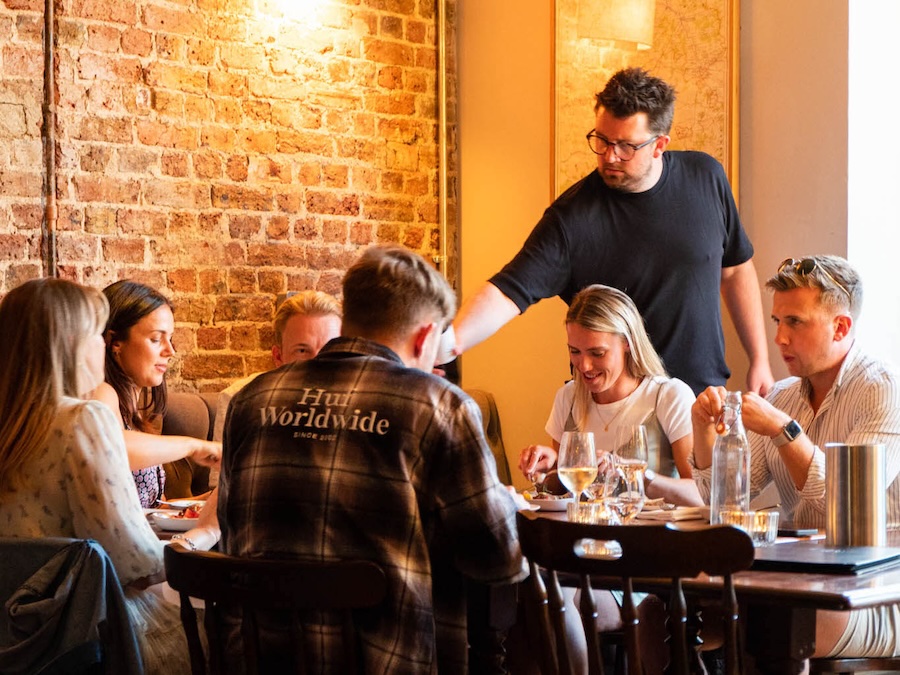 6 people sitting round a dining table in a restaurant. A man in a black t-shirt is leaning in and we assume pouring wine