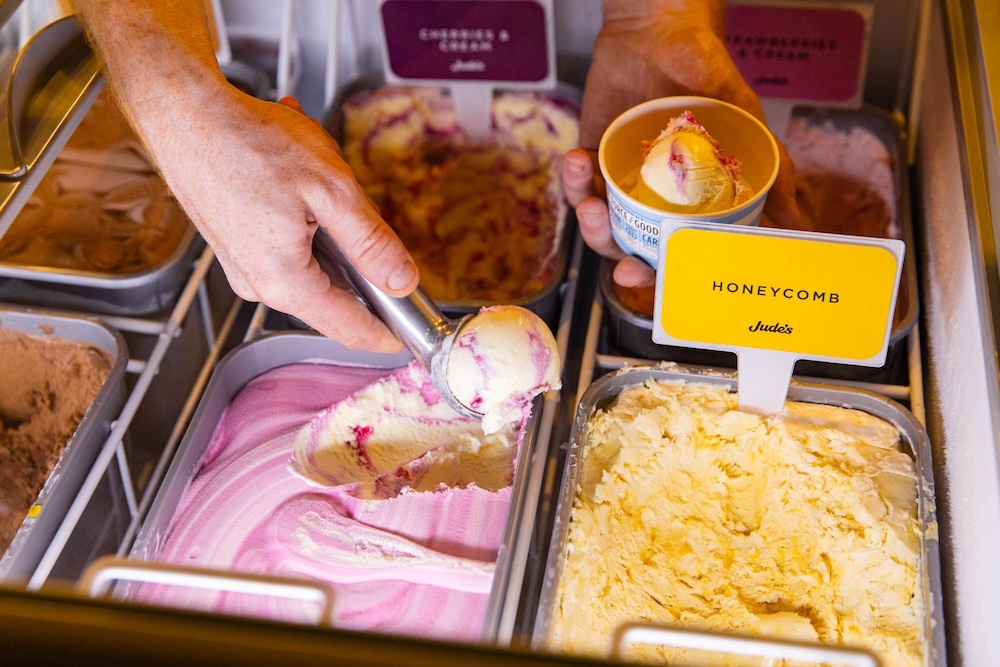 a hand scooping ice cream into a paper cup. Lots of large tubs of ice cream, We see a sign that says honeycomb