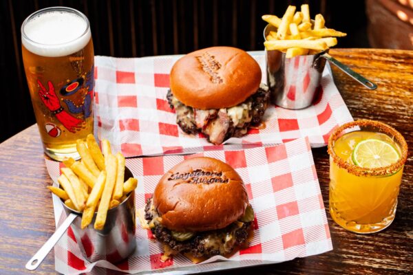 Two burgers with Boysterous Burgers printed on the buns opposite each other on red and white chequered paper. Chips in silver pots. a Pint and a cocktail