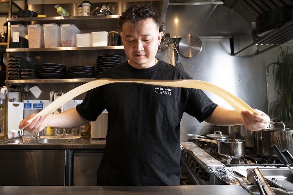a man in a black t-shirt in a kitch stretching out some noodle dough (if that is the right word) between his hands